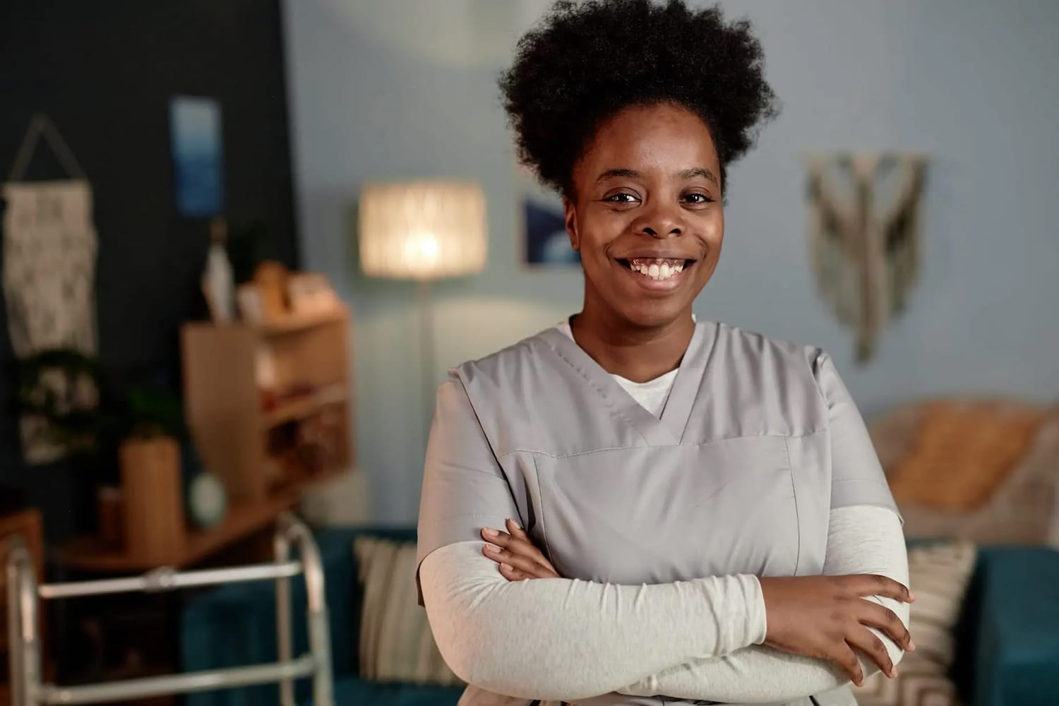 Medium portrait of African American female social worker beaming with smile wearing scrubs posing with arms crossed during house visit to patient, copy space